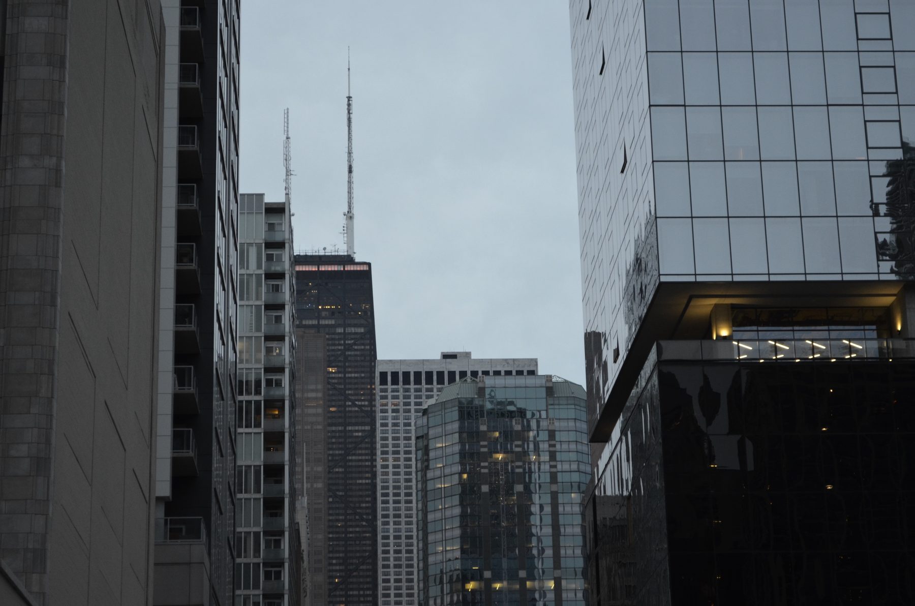 The antennas of the John Hancock Center rising between apartment and glass towers under a gray sky